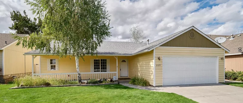 From street picture of the new location which is a yellow house with white trim, short white fence, grass yard, with a tree